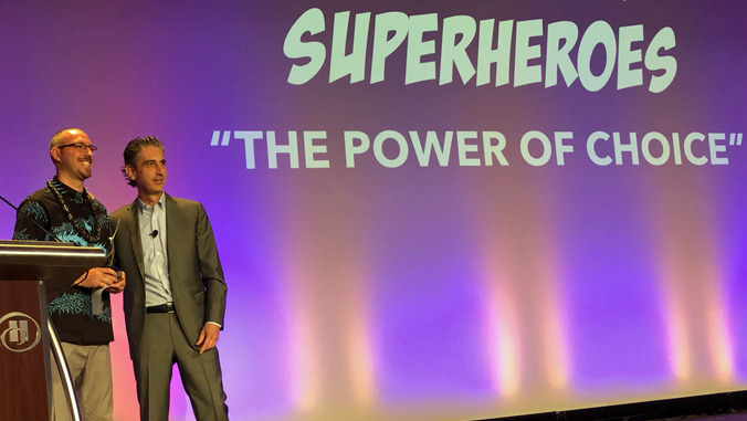 two people standing on a stage with the word Superheroes "The Power of Choice" behind them
