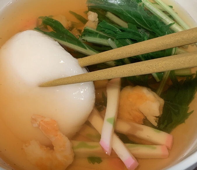 Chopsticks holding a mochi in soup with vegetables and shrimp