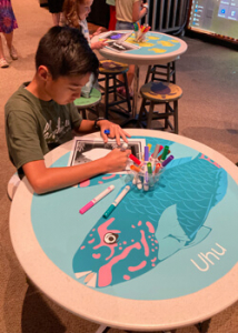 Keiki coloring a paper on a table decorated with the image of an uhu